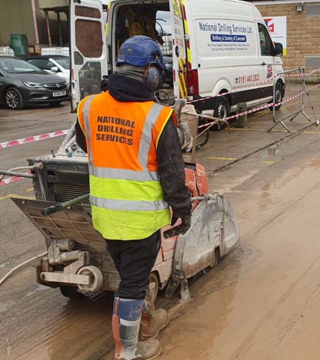 Worker in high-visibility vest with "National Drilling Services" logo operating a concrete floor saw on a construction site, with a company van in the background.